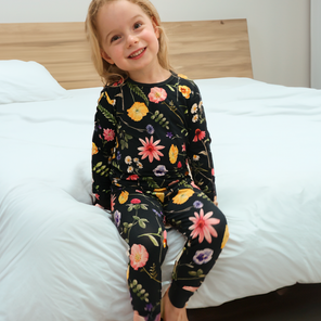 Een kind zit op een bed en draagt een zwarte pyjamaset met lange mouwen en een kleurrijk bloemenpatroon.